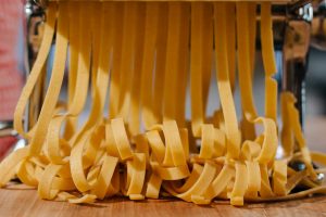 Close-up of fresh pasta strands being made with a pasta machine in an indoor kitchen setting.