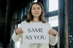 A woman in a white outfit holds a 'Same as You' sign indoors, emphasizing equality.