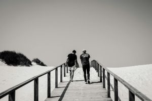 Two men walk on a wooden boardwalk in a snowy beach setting, creating a serene grayscale scene.