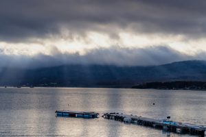 Sunbeams pierce dramatic clouds over a serene lake with a dock in view.