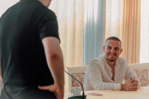 Two men in a casual business meeting setting. One is smiling while seated, communicating friendly interaction.