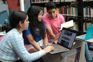 Group of young students studying and discussing in a library setting with books and laptops.