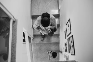 A mother carrying a baby descends a staircase with a young child in a modern home.