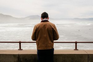 A person stands at a seaside wall, gazing at the misty ocean view under overcast skies.