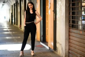 A fashionable woman in a black jumpsuit poses confidently against an urban backdrop.