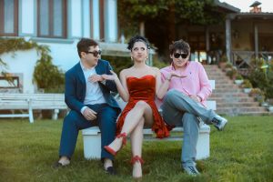 Three young adults in stylish attire sitting together on a bench outdoors, showing camaraderie and elegance.