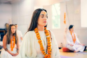A group of women meditating in a tranquil indoor environment, wearing traditional clothing and flower garlands.