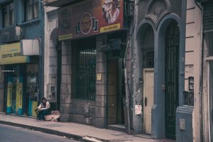 A person sits outside on an urban street near old buildings and shops.
