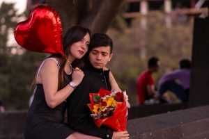 A couple sits close with a red heart-shaped balloon and a bouquet, capturing romance.