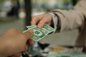 Close-up of hands exchanging Monopoly money outdoors in a park setting.
