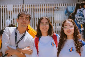 A group of three students smiling outside, wearing uniforms and glasses, embracing camaraderie.