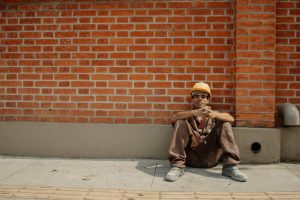 Tired construction worker sitting against brick wall in sunny urban setting.