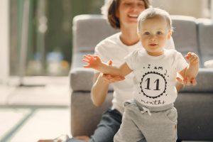 Adorable infant boy looking at camera while walking with support of loving mother in well lit living room