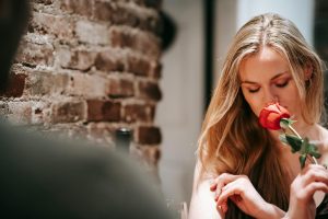 Young couple in elegant clothes in light cafeteria near brick wall having romantic dinner while smelling red rose