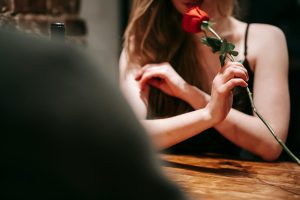 Crop faceless young couple in elegant outfit sitting at wooden table in restaurant and enjoying date with red rose