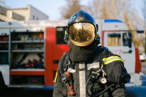 Firefighter in full protective gear with a reflective helmet stands in front of a fire truck on a sunny day.
