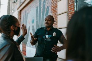 Police officer conversing with people on the street near a brick wall mural.