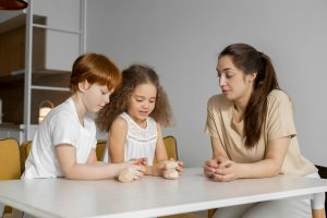 Children and woman sitting together at a white table, playing with wooden blocks.