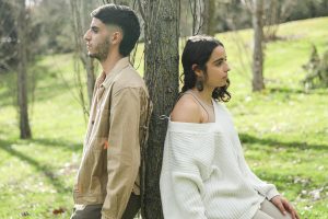 A young man and woman sit back to back against a tree in a sunny park.