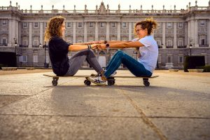 Two women sitting on skateboards and holding hands in front of a historical building, showcasing friendship and fun.