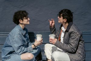 Side view of young stylish ladies with short dark hairs in casual outfits sitting on wooden bench with crossed legs and chatting while drinking takeaway coffee during weekend on street