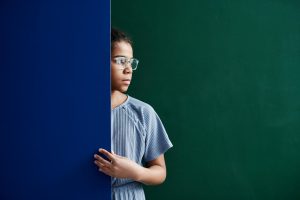 Teen girl wearing eyeglasses stands by a blue partition, gazing thoughtfully.