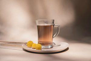 A clear tea glass with brown tea and yellow flowers on a white saucer in soft lighting.