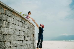 Side view of Asian male in sportswear standing on picturesque sandy beach and stretching arms towards child standing on waterfront on clear summer sunny day