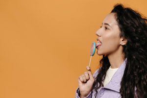 Portrait of a woman licking a colorful lollipop against a bright orange backdrop, offering ample copy space.