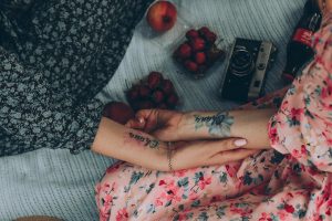 Two individuals holding hands on a picnic blanket with fruits and vintage camera, showcasing floral dresses and tattoos.