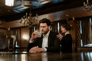 An elegant bar scene with a thoughtful man holding a glass and a woman comforting him.