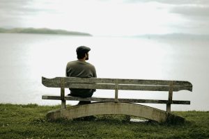 A man in a flat cap sits on a wooden bench, enjoying a peaceful lakeside view.