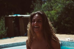 Woman smiling joyfully near a swimming pool on a sunny day, enjoying nature.