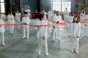 A diverse group of individuals in white clothing connected by a red ribbon indoors, symbolizing unity and diversity.