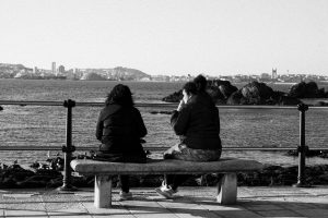 Black and white photo of two women on a bench with a scenic city and sea view.