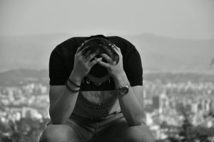 A focused young man sits with hands on head against an urban cityscape in grayscale, suggesting introspection.