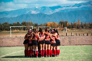 A women's field hockey team gathers in a huddle on a field with mountains in the background during fall.