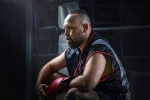 Bearded male boxer in sportswear sitting in boxing ring, contemplating during training session.