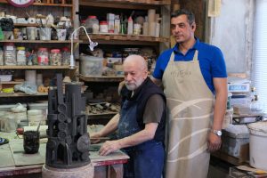 Elderly sculptor and younger apprentice working on pottery in an artisan's studio.