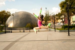 A young female dancer leaping joyfully in front of a modern dome structure in a vibrant city setting.