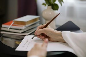 A close-up of a person writing with a fountain pen on paper, books stacked nearby indoors.