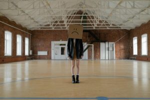 A person wearing a cardboard box with a sad face in an empty gym, evoking melancholy.