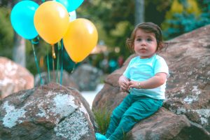 Charming toddler seated on rocks with colorful balloons in a tranquil park.