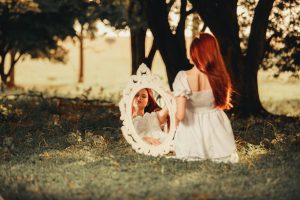 A redhead woman sits outdoors with a mirror reflection, surrounded by nature.