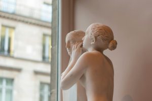 A delicate marble sculpture depicting two figures in a whispering pose by a glass window.