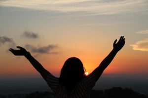 Silhouette of a woman with raised arms embracing the sunset scenery from a hilltop view.
