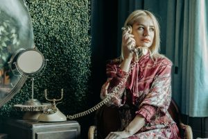 A woman in vintage attire uses a rotary phone, evoking retro elegance indoors.