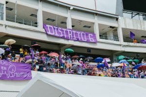 A lively crowd in a stadium stands under umbrellas during a community event on a bright day.