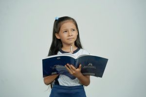 Young schoolgirl with long hair holding a book and pencil, deep in thought against a plain background.