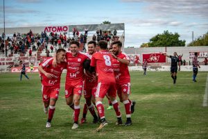 Group of soccer players in red uniforms celebrating a goal on an outdoor football field.